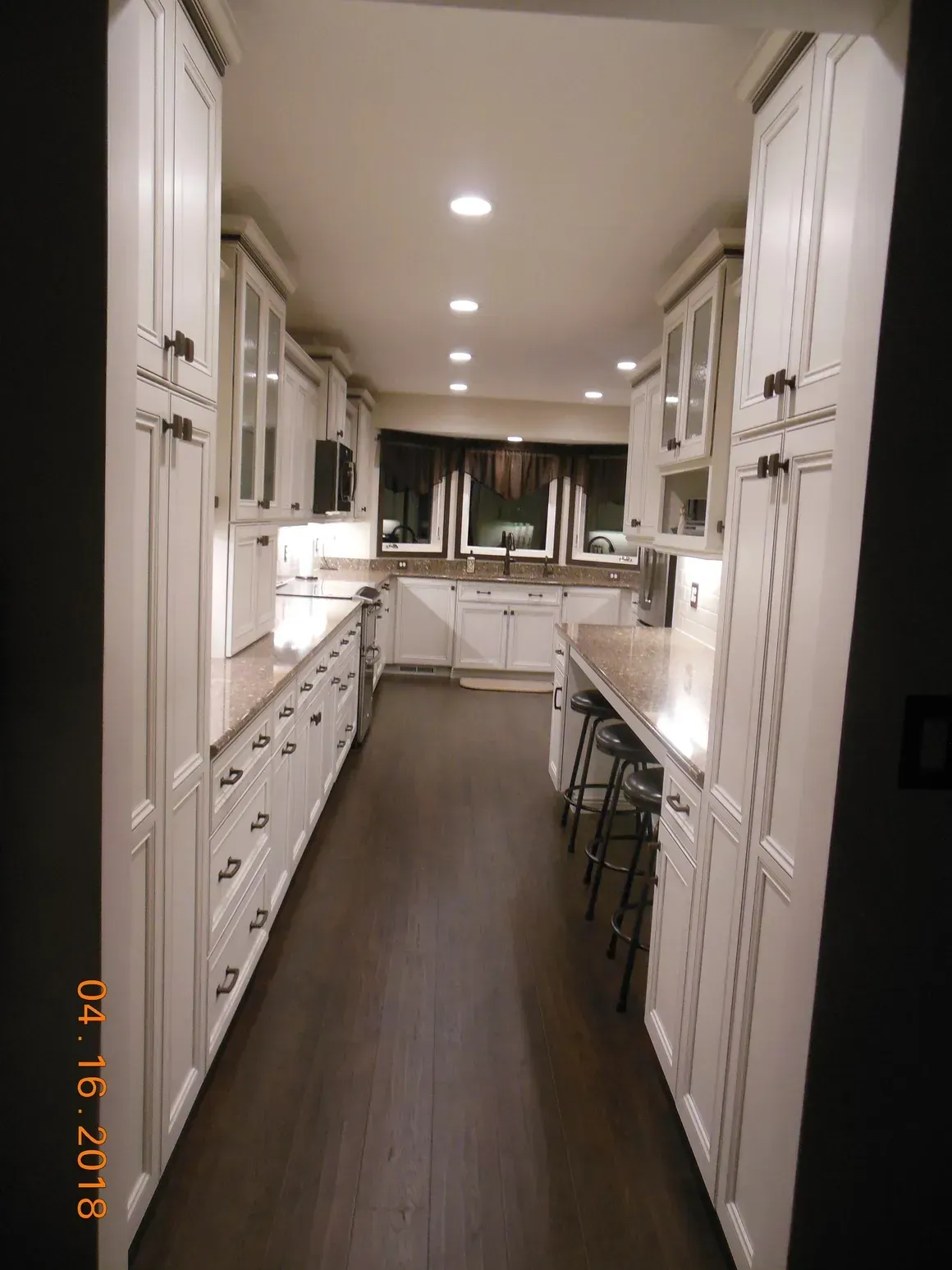 A narrow galley kitchen with white cabinets, light countertops, dark wood flooring, and recessed ceiling lights.