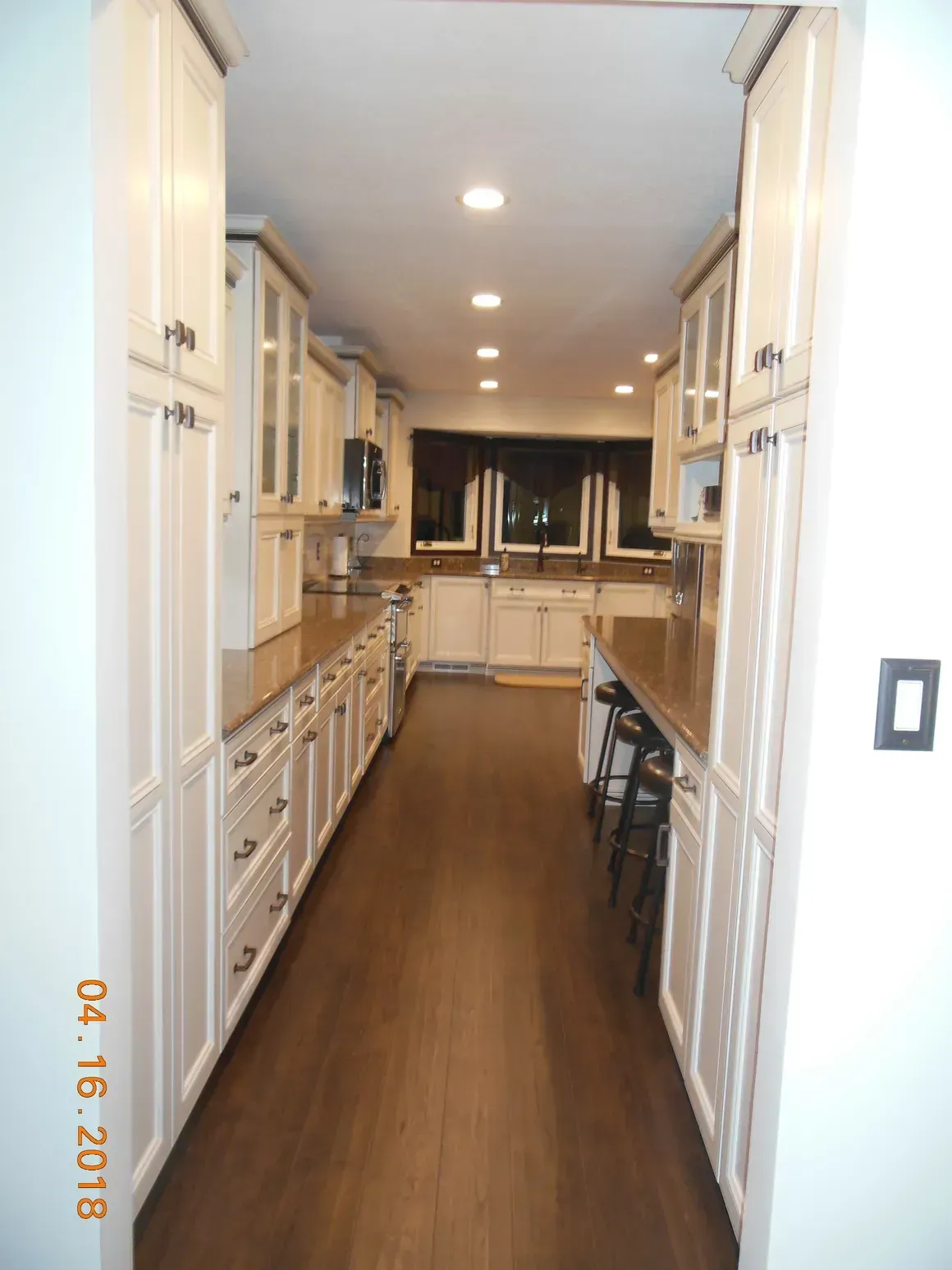 A long galley kitchen with cream-colored cabinets, brown countertops, and wood flooring, seen from a doorway.