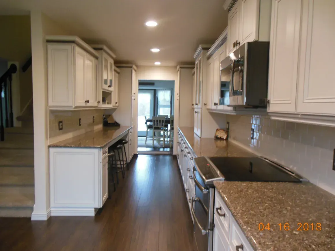 Galley kitchen with white cabinets, brown granite countertops, dark wood flooring, and a view of a dining area.