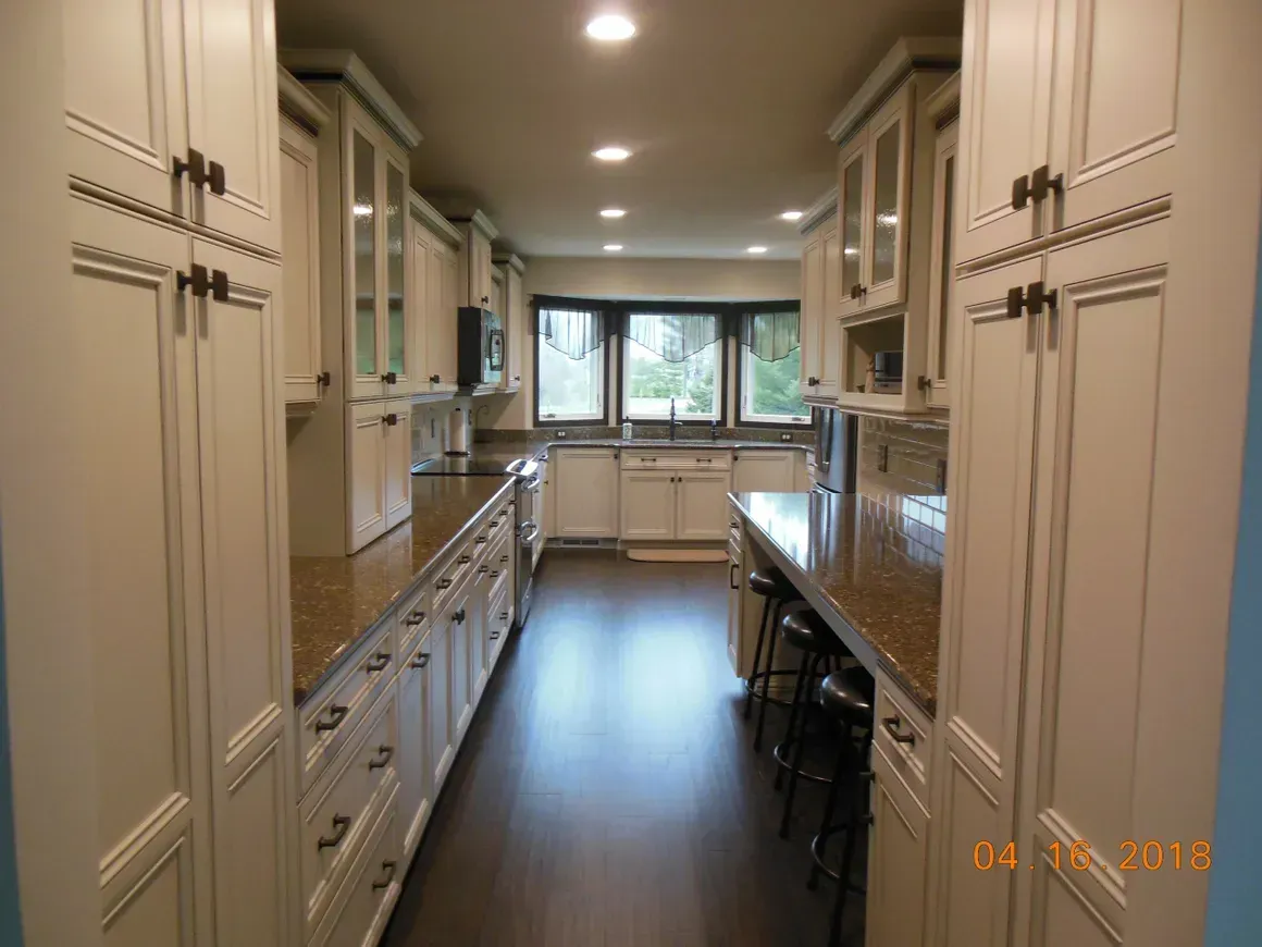 A galley-style kitchen with cream-colored cabinetry, brown countertops, dark wood flooring, and a window at the end.
