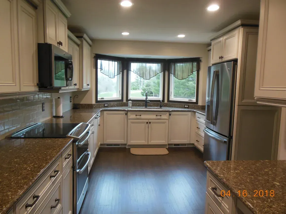 A kitchen with white cabinets, dark countertops, stainless steel appliances, and a window above a central sink.