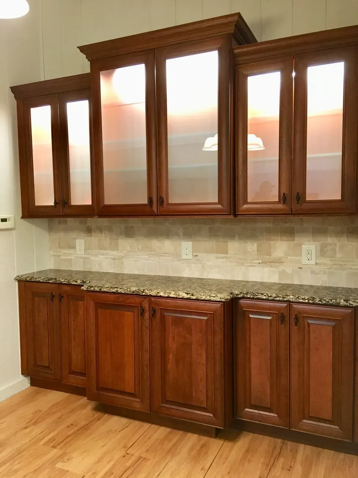 A wall of dark wood cabinets with frosted glass upper doors, granite countertops, and stone backsplash in a kitchen.