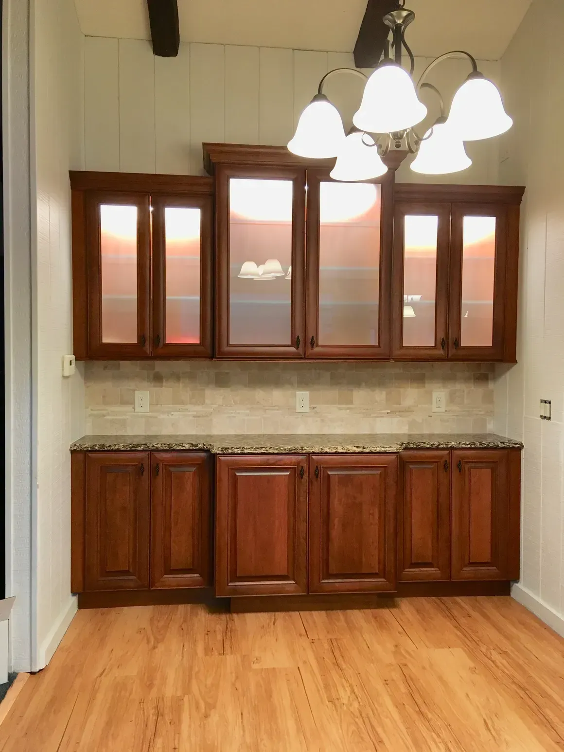 A wooden built-in buffet with glass-front upper cabinets, a tiled backsplash, and a hanging chandelier in a dining room.