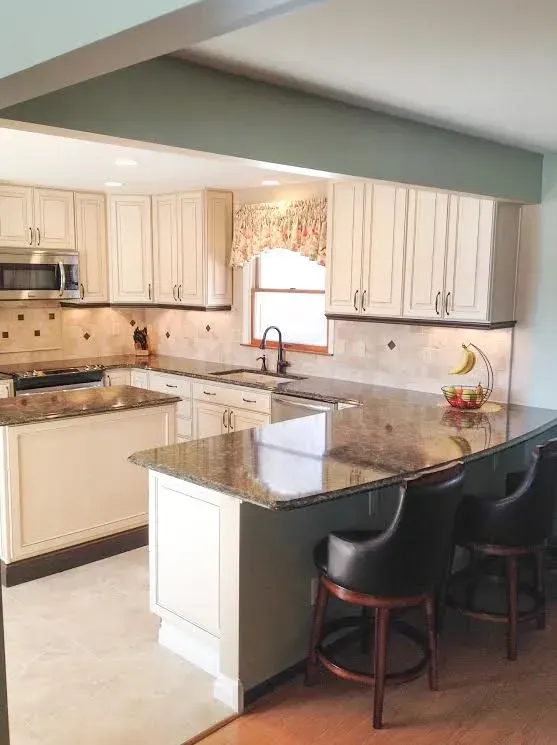A brightly lit kitchen with cream-colored cabinets, dark granite countertops, and two black bar stools at the island.