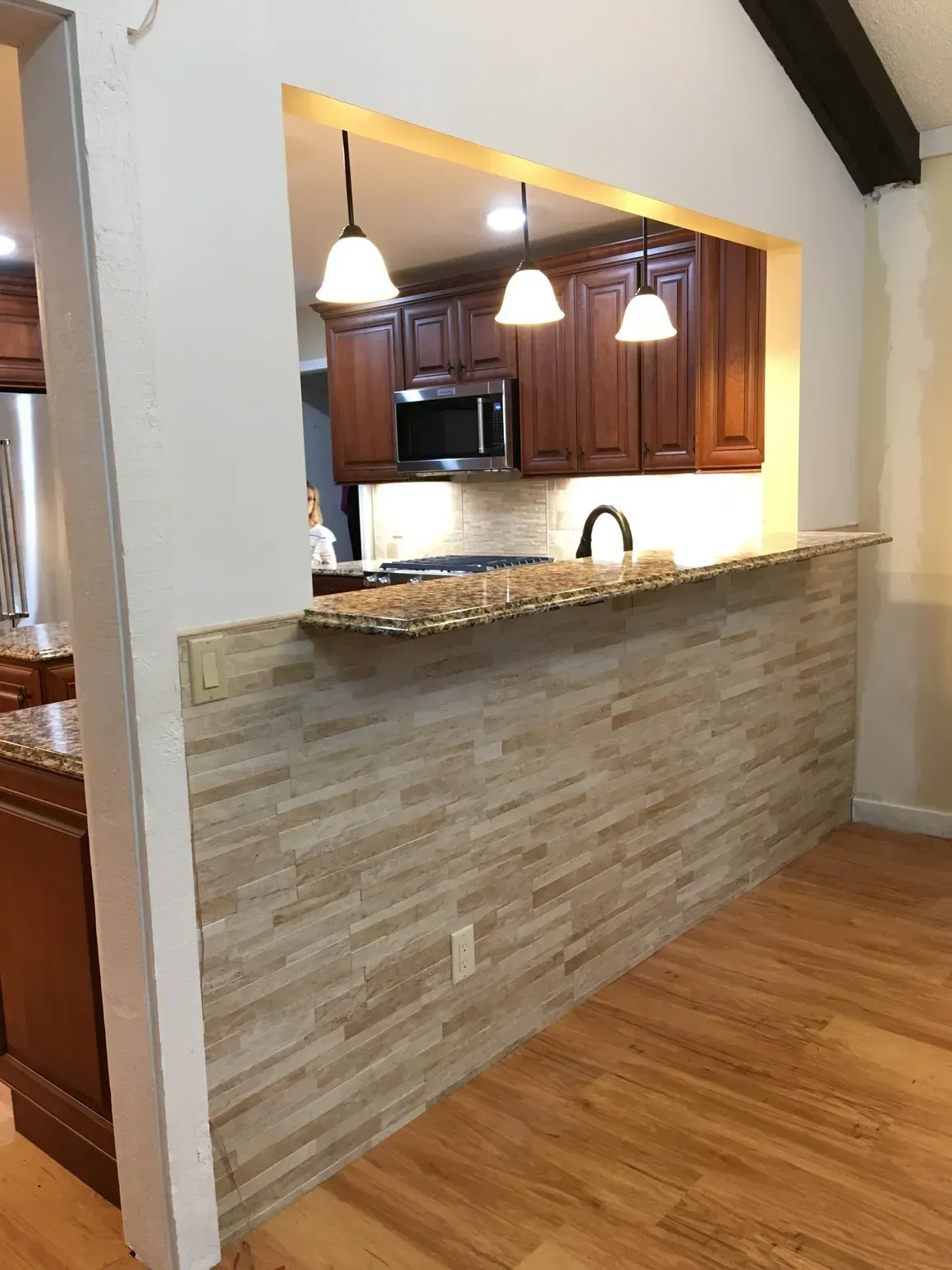 A kitchen pass-through with a stone-tiled half wall, granite countertop, pendant lighting, and warm-toned wood cabinets.