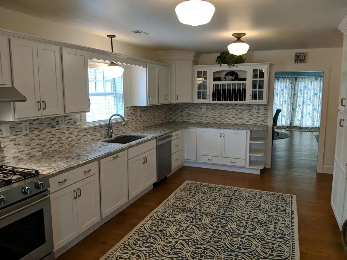 A bright kitchen features white cabinets, stone countertops, a patterned rug, and natural light from a window.