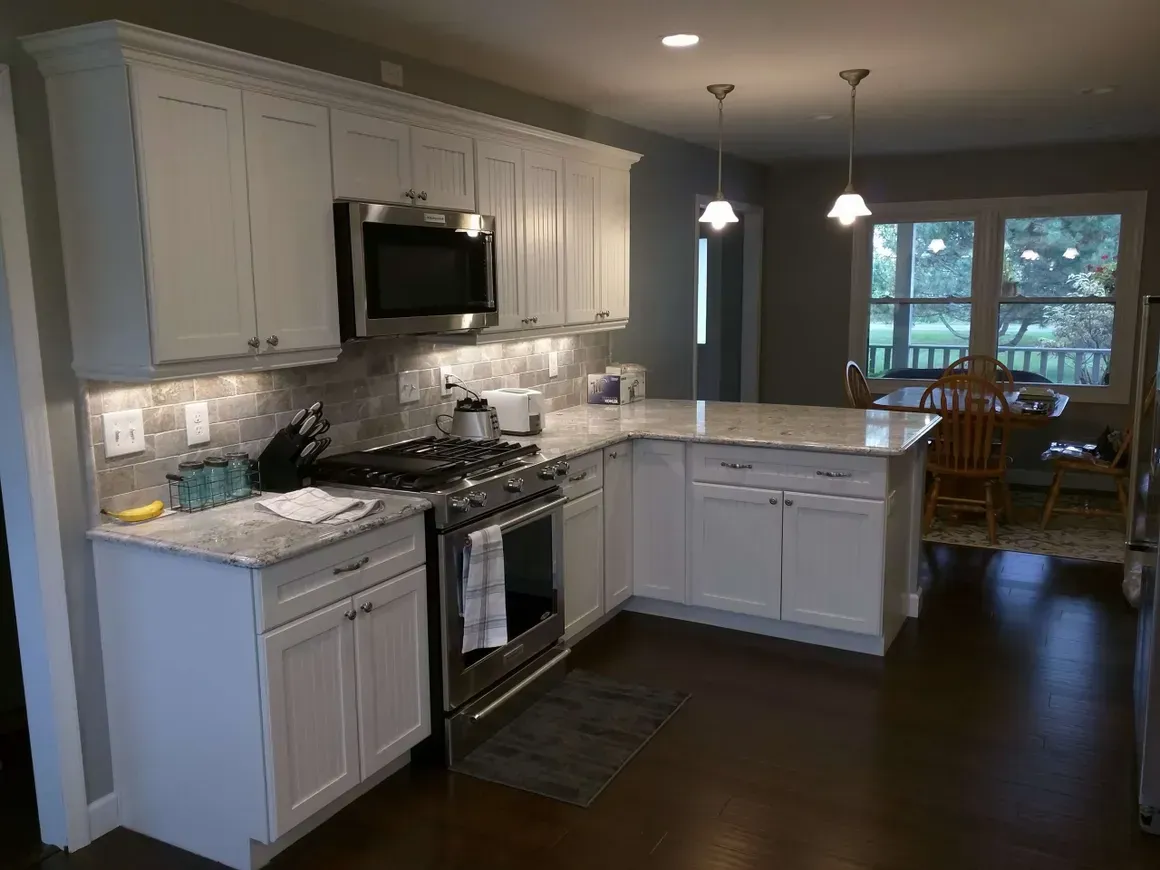 A modern white kitchen featuring light granite countertops, stainless steel appliances, and a dining area in the back.