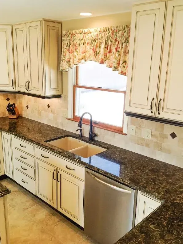 Kitchen with cream-colored cabinets, dark granite countertops, a double sink under a window, and a stainless dishwasher.