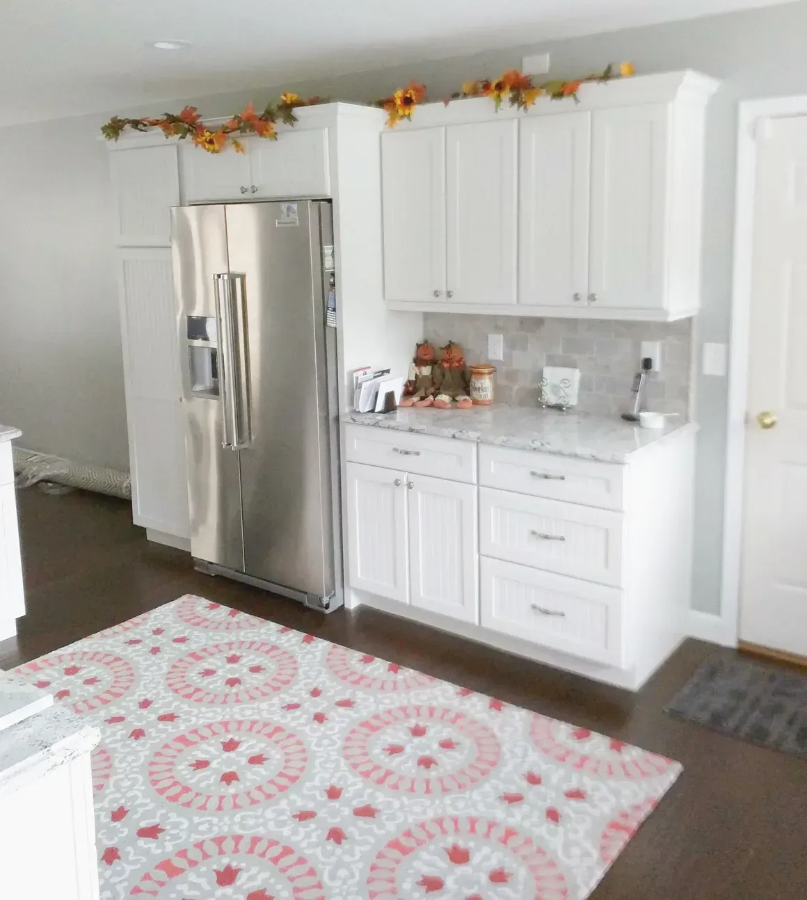 White kitchen cabinets, a stainless steel refrigerator, and a patterned rug on a dark wood floor.