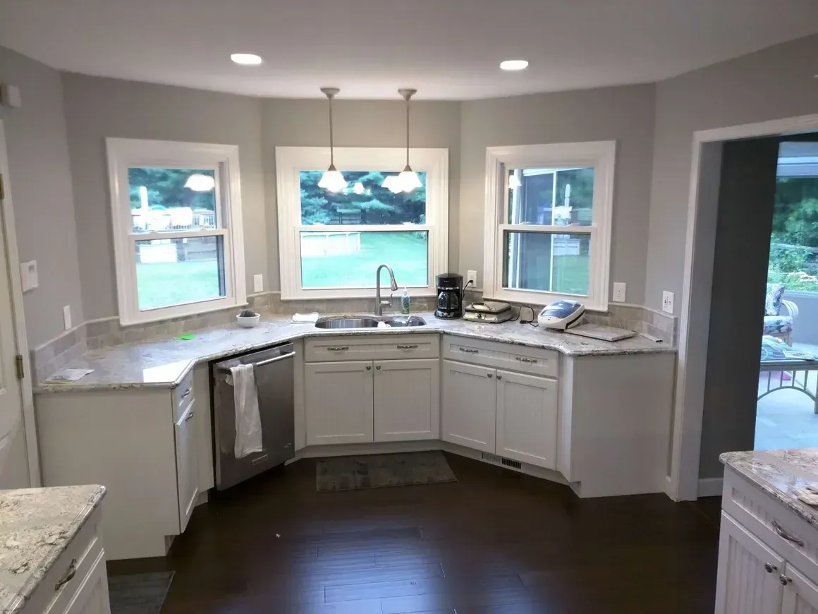 A kitchen featuring white cabinets, granite countertops, three windows above a corner sink, and dark wood flooring.