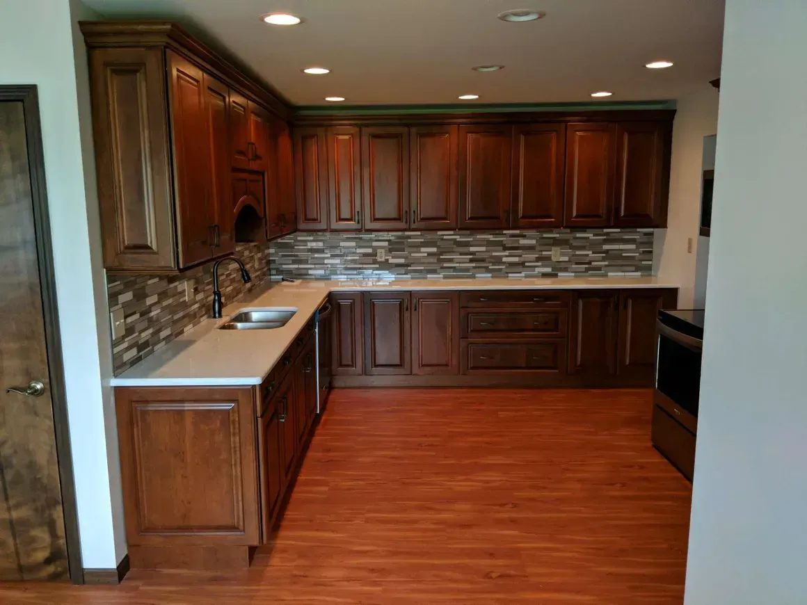 A modern kitchen with dark wood cabinets, a light stone backsplash, cream-colored countertops, and reddish-brown flooring.