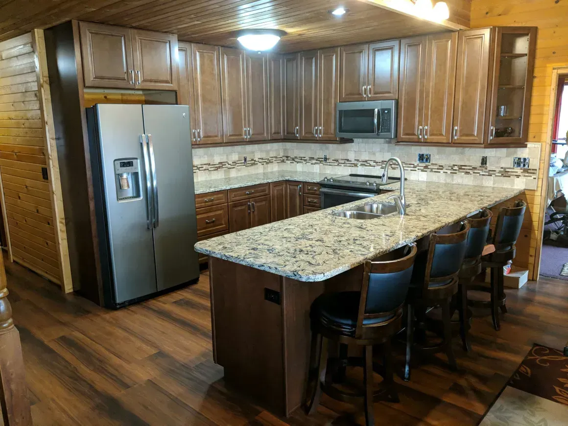 A rustic, L-shaped kitchen featuring wooden cabinets, granite countertops, a stainless steel refrigerator, and bar stools.