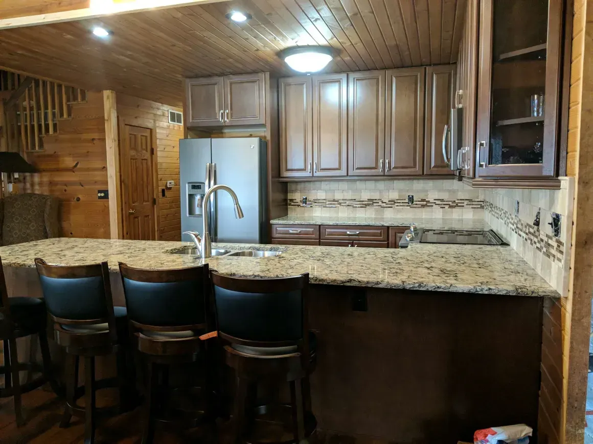 A rustic kitchen with wood-paneled walls and ceilings, featuring dark wood cabinets, a granite island, and bar stools.