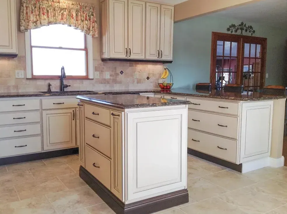 A kitchen featuring cream-colored cabinets, a granite-topped central island, a tile backsplash, and light-colored tile floors.