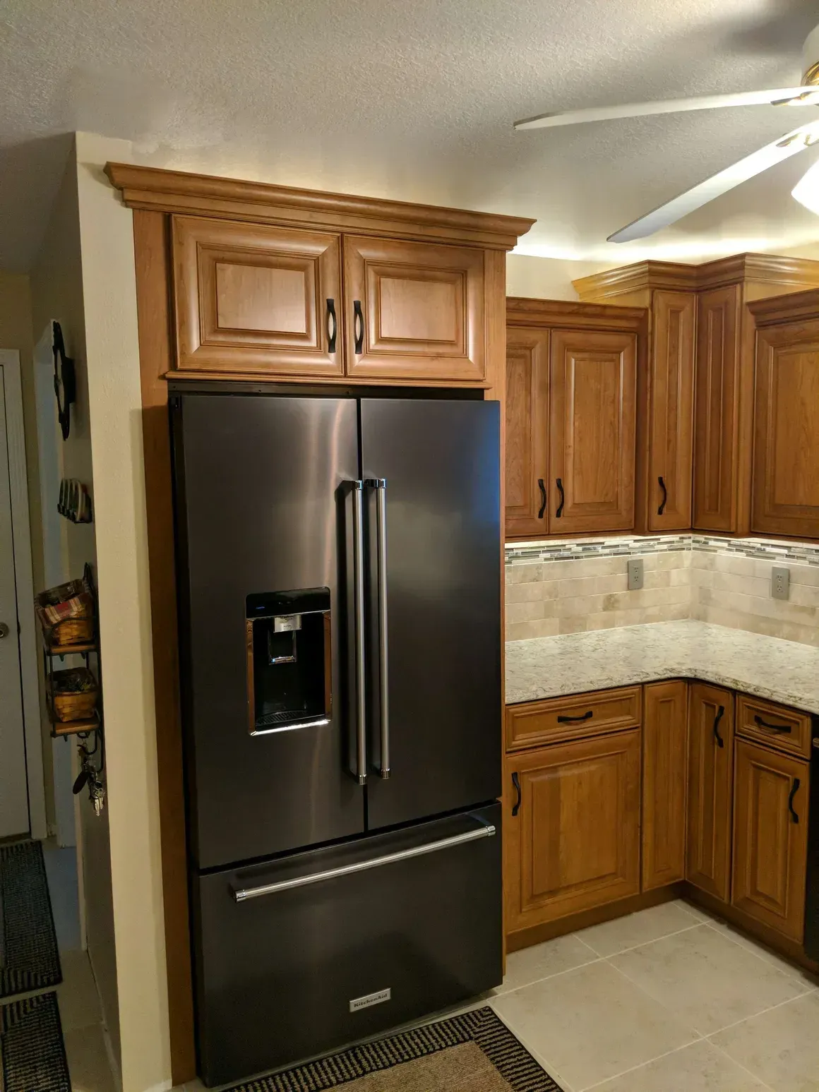 A dark stainless steel refrigerator sits in a kitchen cabinet alcove with oak cabinetry and light-colored tiled counters.