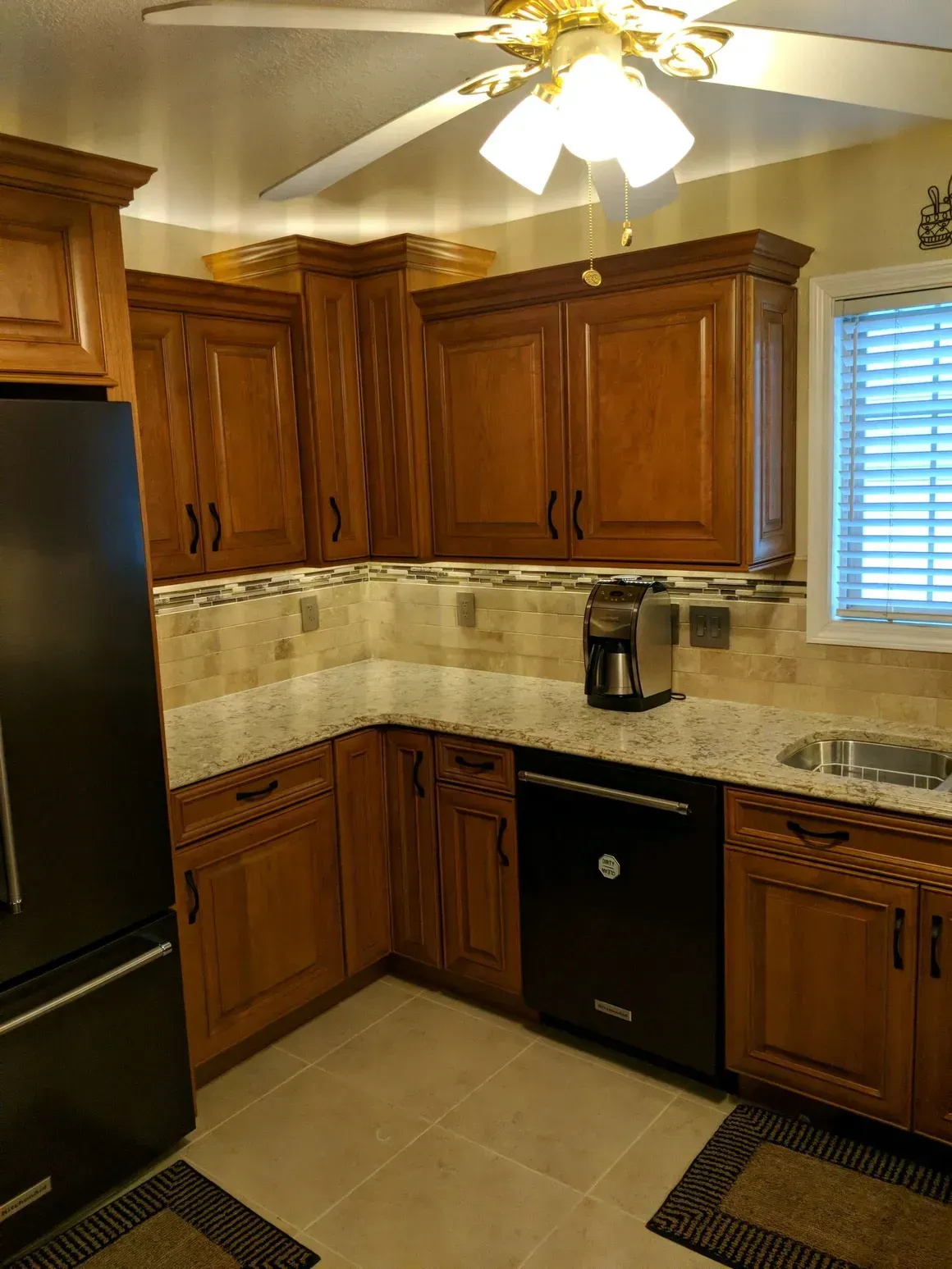A kitchen with wood cabinets, light granite countertops, a black dishwasher, and a ceiling fan over a tiled floor.