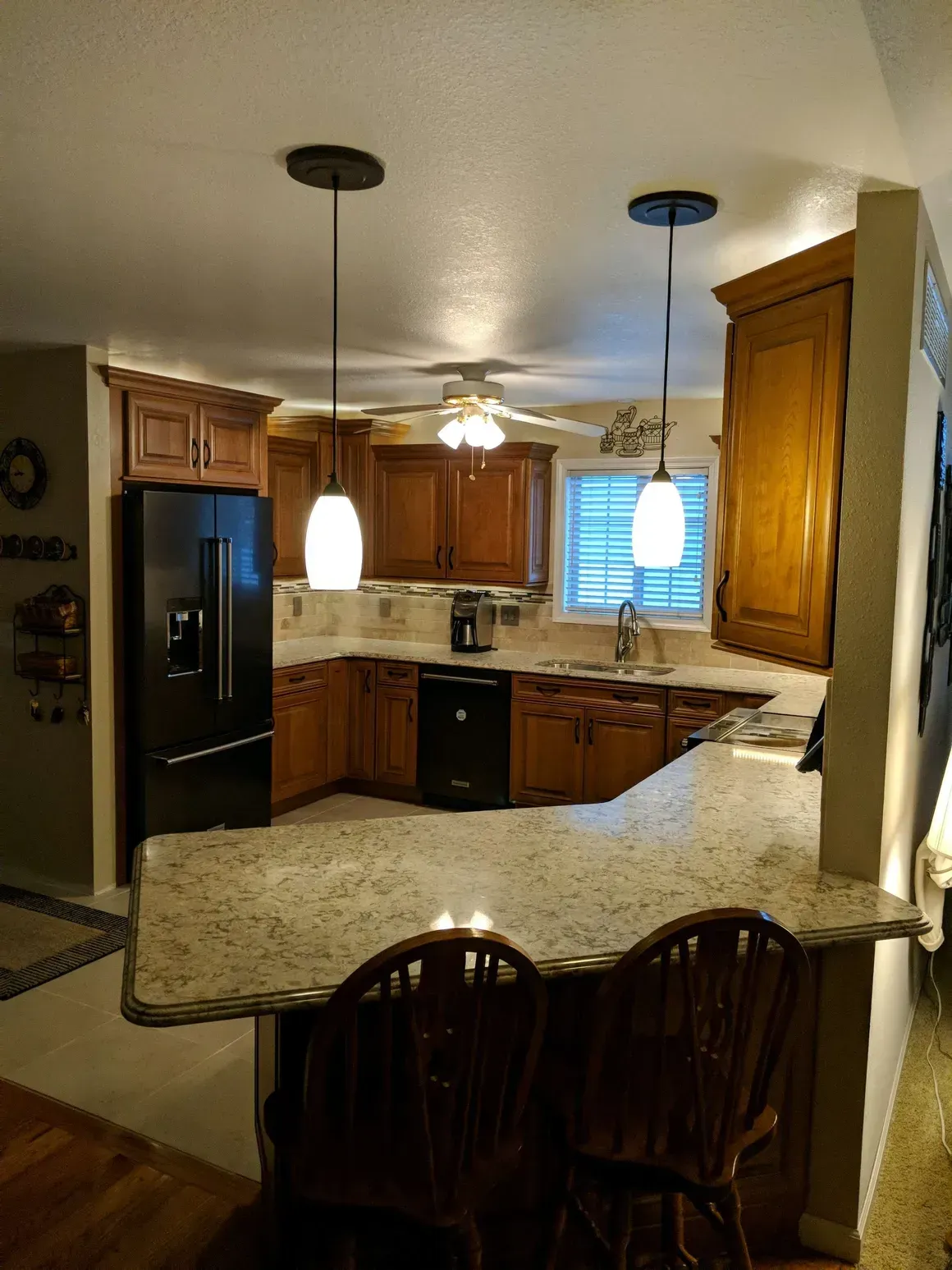 Kitchen with wood cabinets, black appliances, a speckled granite countertop island with two chairs, and two pendant lights.