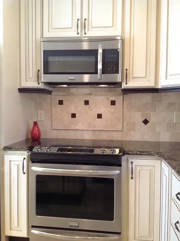 Stainless steel oven and microwave in a kitchen with off-white cabinets, dark countertops, and a decorative tile backsplash.