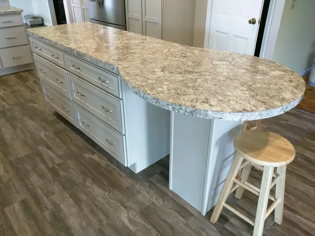 A kitchen island with a beige speckled countertop, white cabinets, and a single wooden stool on a wood-look floor.