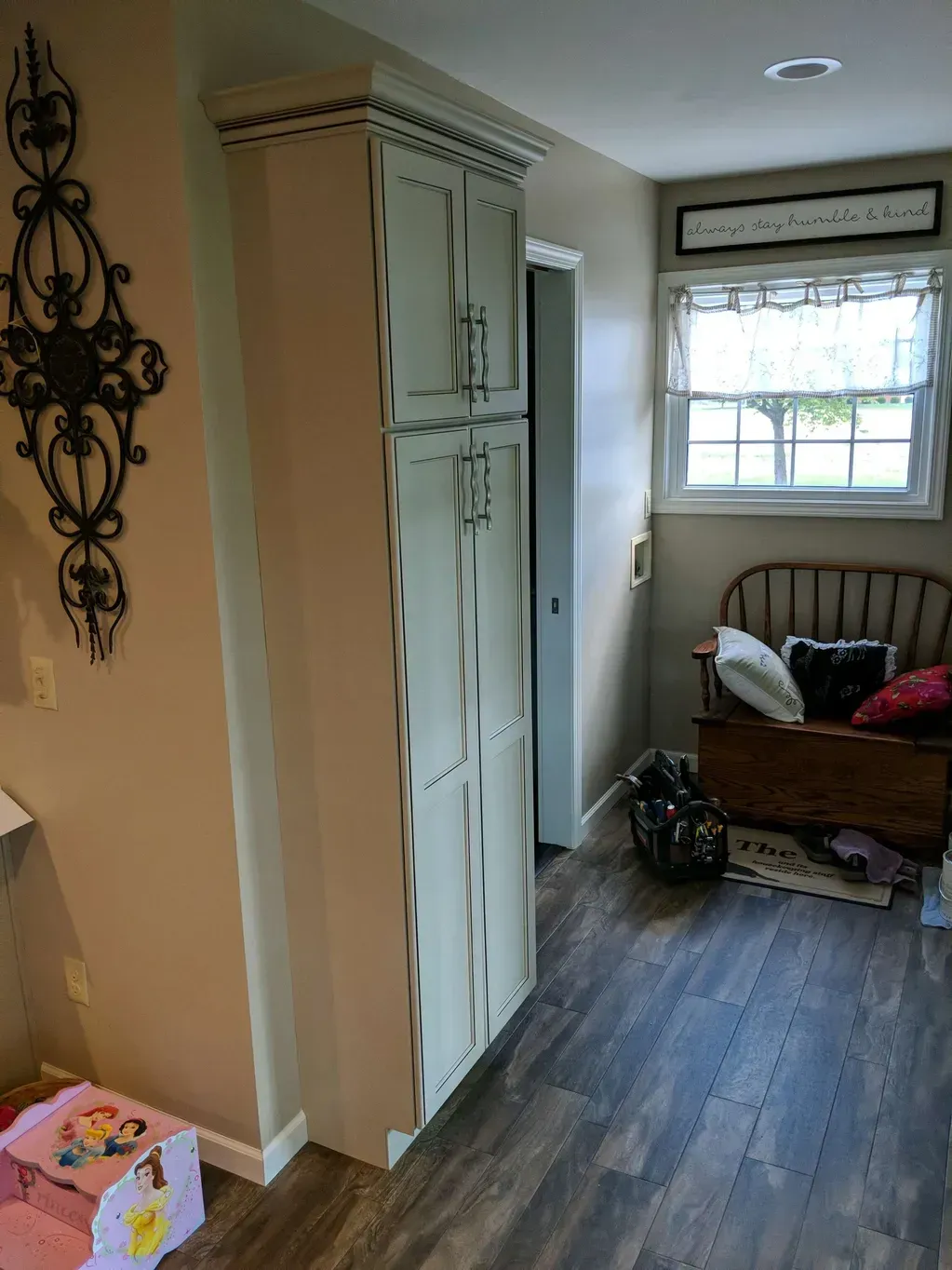 A tall, distressed cream cabinet sits next to a doorway in a hallway with wood flooring and a wooden bench by the window.
