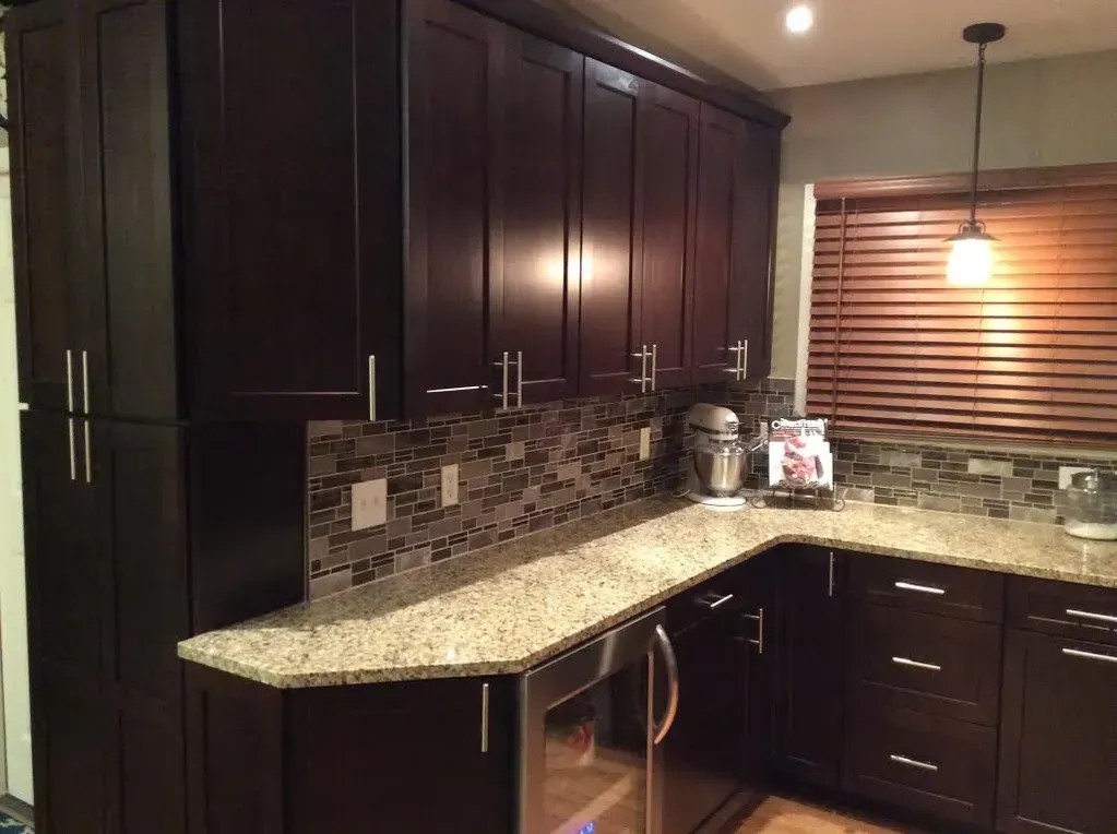 A kitchen with dark wood cabinets, speckled granite countertops, a mosaic tile backsplash, and a pendant light.