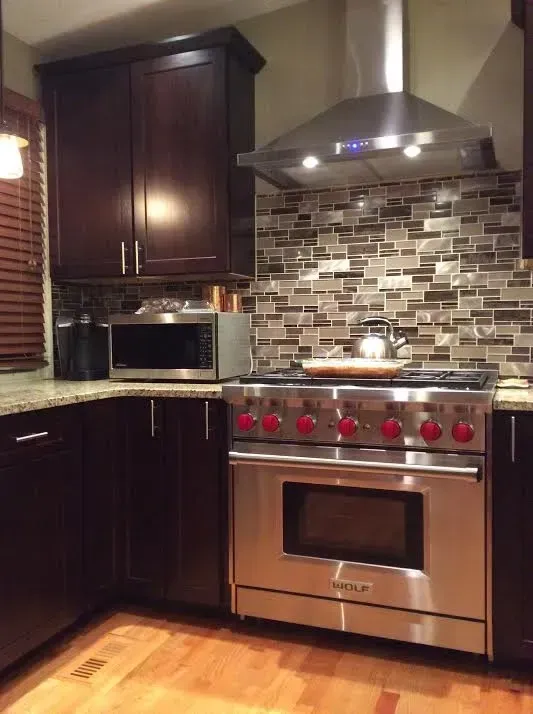 Kitchen featuring dark cabinets, a stainless steel stove with red knobs, a range hood, and a multi-colored tile backsplash.
