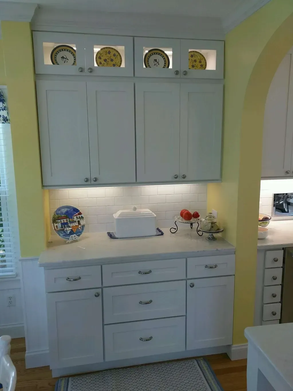 White kitchen cabinetry featuring glass-front display shelves, drawers, and a white countertop with a decorative plate.