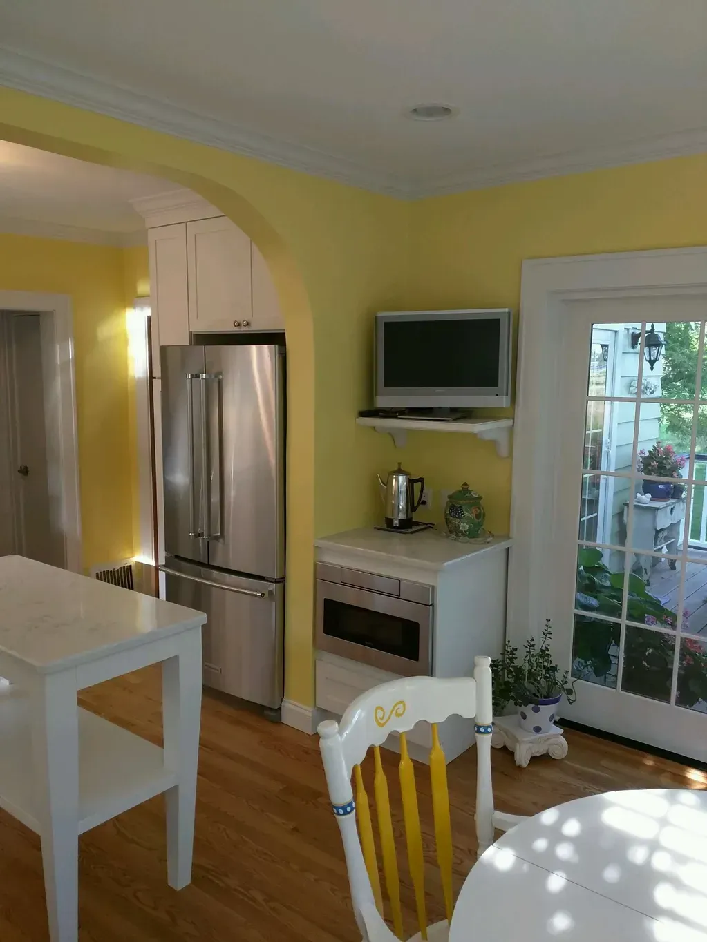 A bright kitchen with yellow walls, a stainless steel refrigerator, white cabinetry, and a small television on a shelf.