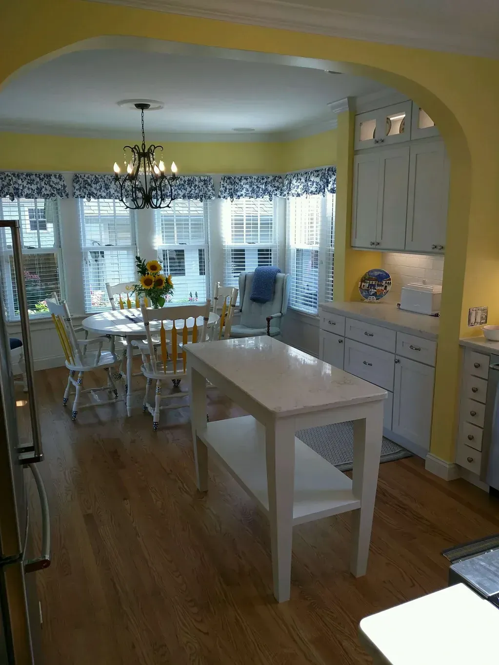 A bright yellow kitchen features a white island in the foreground and a dining table with chairs under a windowed alcove.