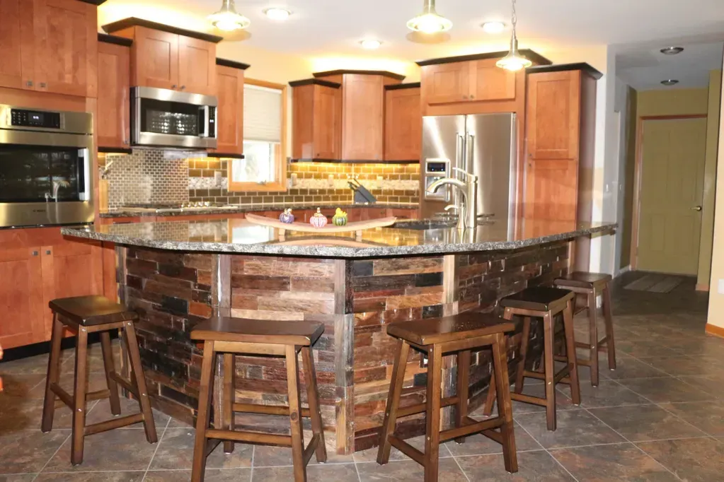 A kitchen island with a dark wood plank exterior, stone countertop, and four wooden stools in a wood-cabinet kitchen.