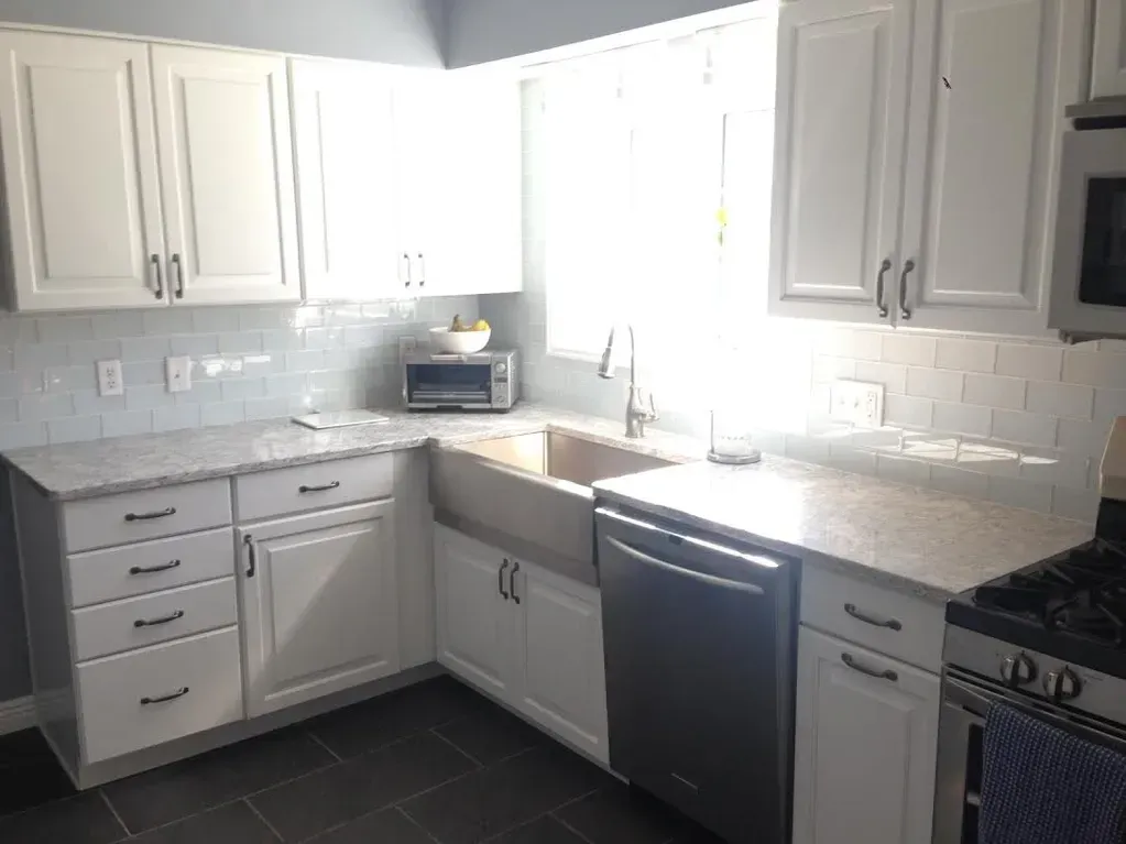 A kitchen featuring white cabinets, light speckled countertops, a farm sink, and grey tile floors.