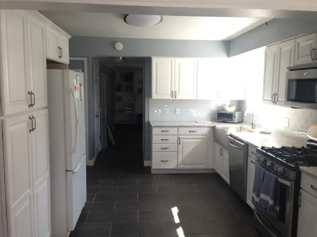 A kitchen with white cabinets, dark tile floors, stainless steel appliances, and an open doorway leading to a dark hallway.