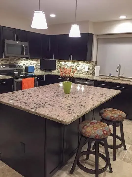 A kitchen island with a granite countertop and two stools stands in front of dark cabinets and a stone backsplash.