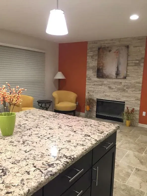 A kitchen island with a speckled granite countertop in front of a stone fireplace with orange walls and yellow chairs.