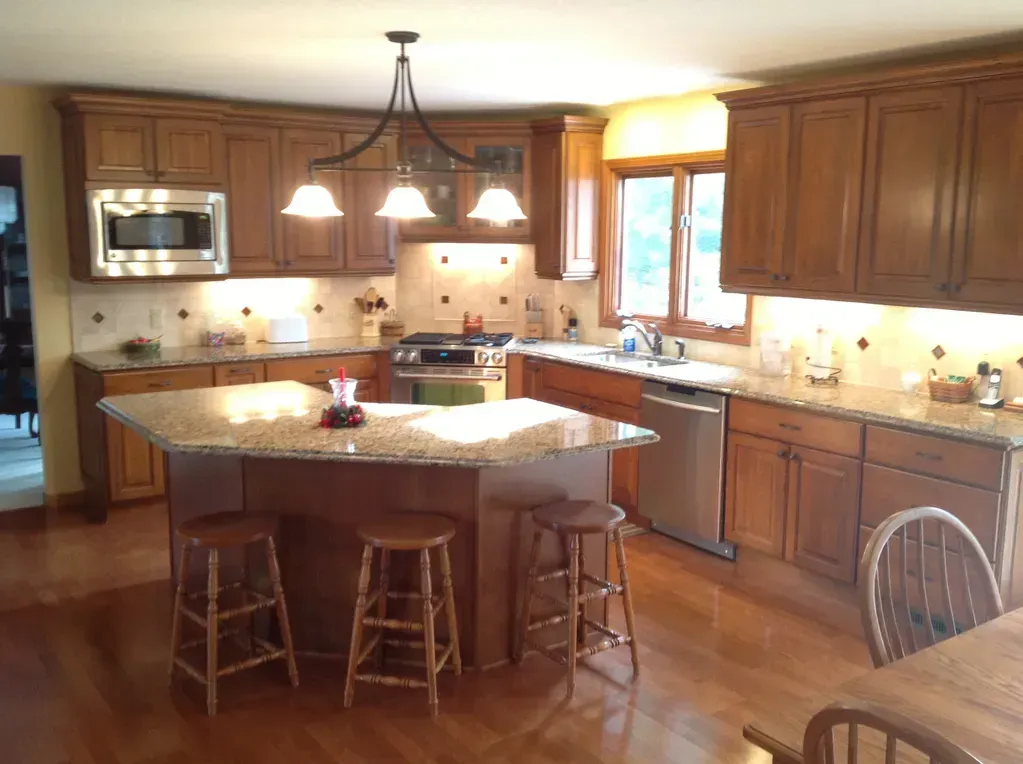 A kitchen with wood cabinets, granite countertops, a central island with three bar stools, and a stainless steel appliance.