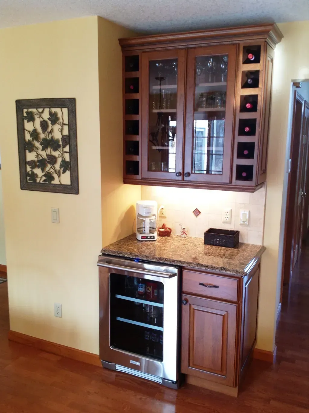A wooden wine bar with a glass-front fridge, granite counter, and wall-mounted cabinet with integrated wine bottle storage.