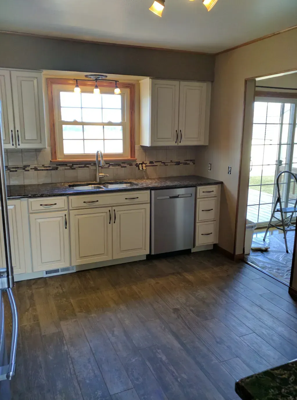 A kitchen with cream-colored cabinets, dark countertops, a stainless steel dishwasher, and a window over the sink.