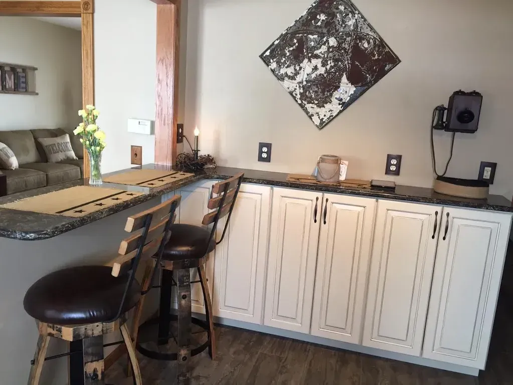 A home bar area featuring white cabinets, a dark countertop, two bar stools, and a diamond-shaped wall decoration.