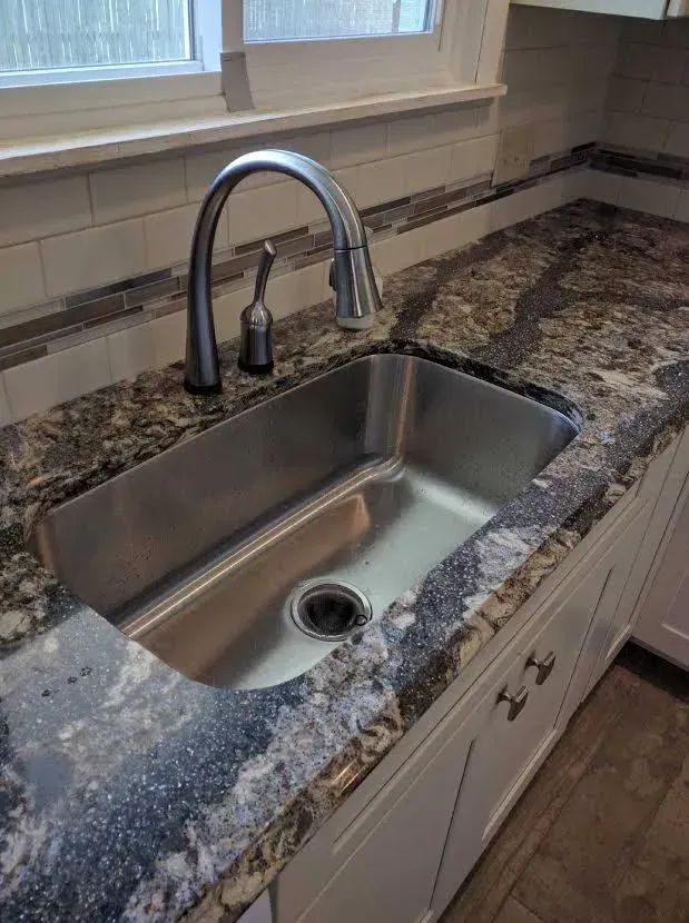 A stainless steel kitchen sink with a high-arch faucet, set into a dark, patterned granite countertop against a tiled wall.