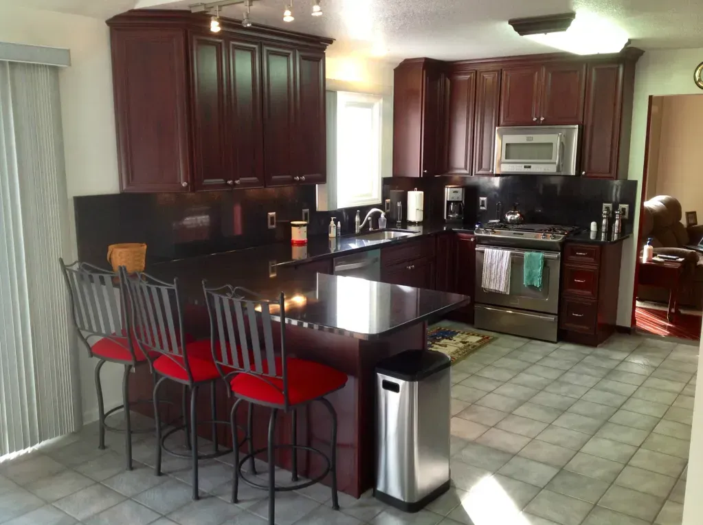 A kitchen with dark wood cabinets, a black island, three red-cushioned bar stools, and stainless steel appliances.