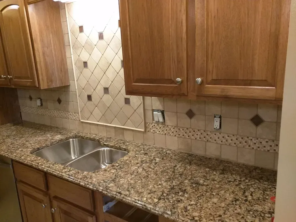 A kitchen sink area featuring wooden cabinets, granite countertops, and decorative tiled backsplashes.