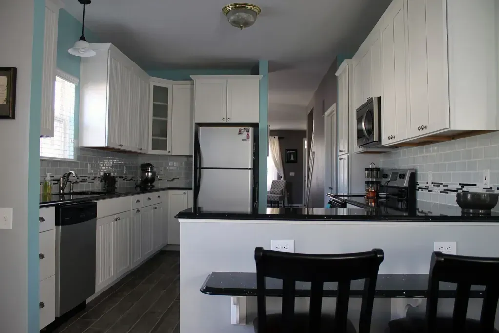 A modern kitchen with white cabinets, stainless steel appliances, a black countertop, and a breakfast bar with two chairs.