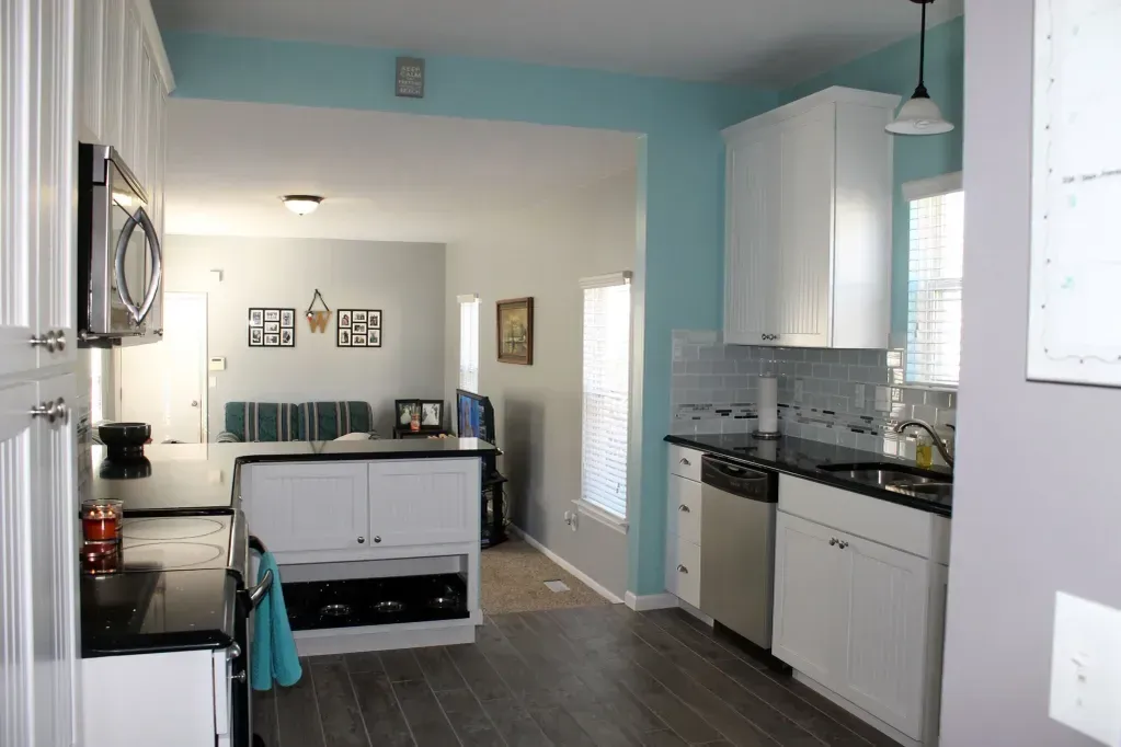 A kitchen with white cabinets, dark countertops, and light blue walls, transitioning into a living room with gray walls.