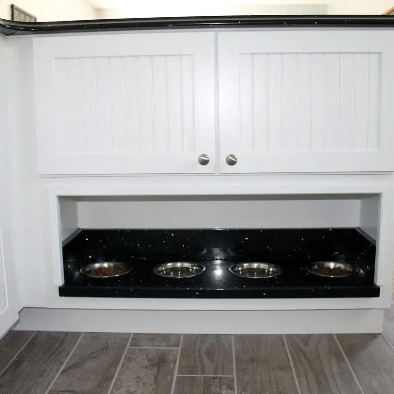 A built-in kitchen cabinet features a low, open shelf with four pet food bowls sitting on a black granite countertop.