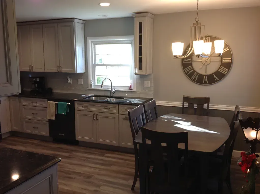 A kitchen with grey cabinets, dark countertops, a window over the sink, and a dining table with chairs under a chandelier.