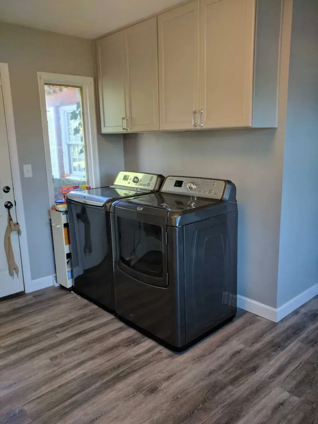 A laundry area with a dark gray washer and dryer beneath white wall cabinets on a light wood-look floor near a door.