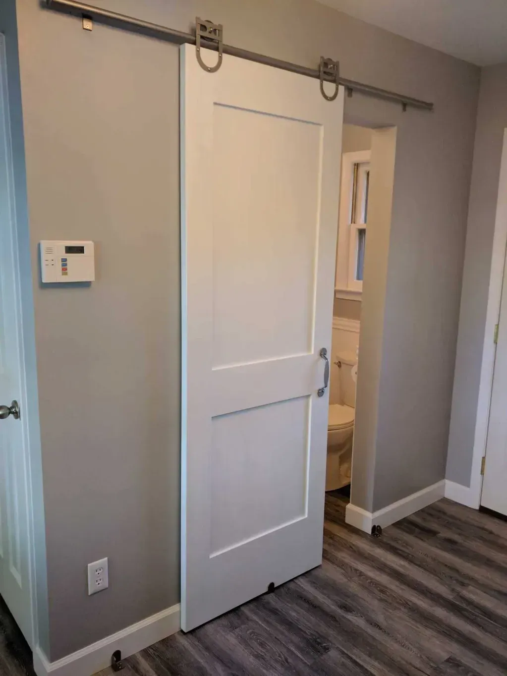 A white sliding barn door on a metal track installed over the doorway of a small bathroom with gray walls and wood floors.