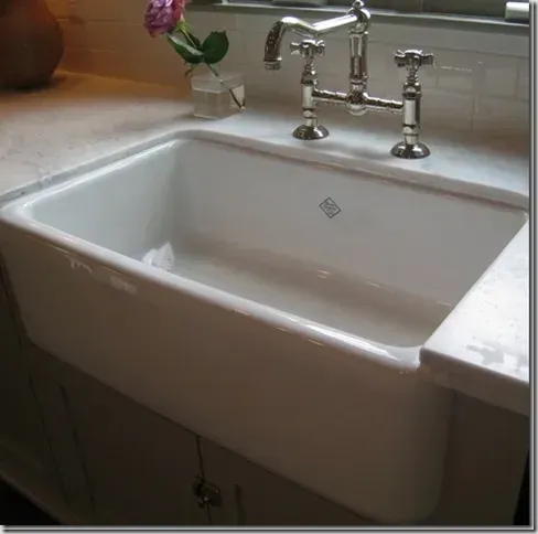 A white farmhouse sink with a chrome bridge faucet, set in a stone countertop with a small flower in a vase.