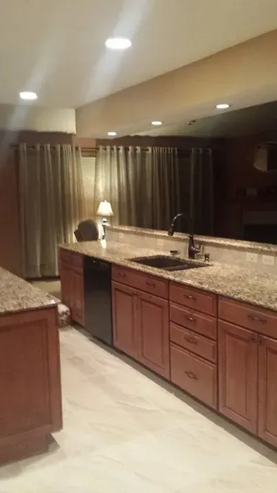 A kitchen with wooden cabinets, granite countertops, a dark dishwasher, and light-colored flooring, with curtains behind.