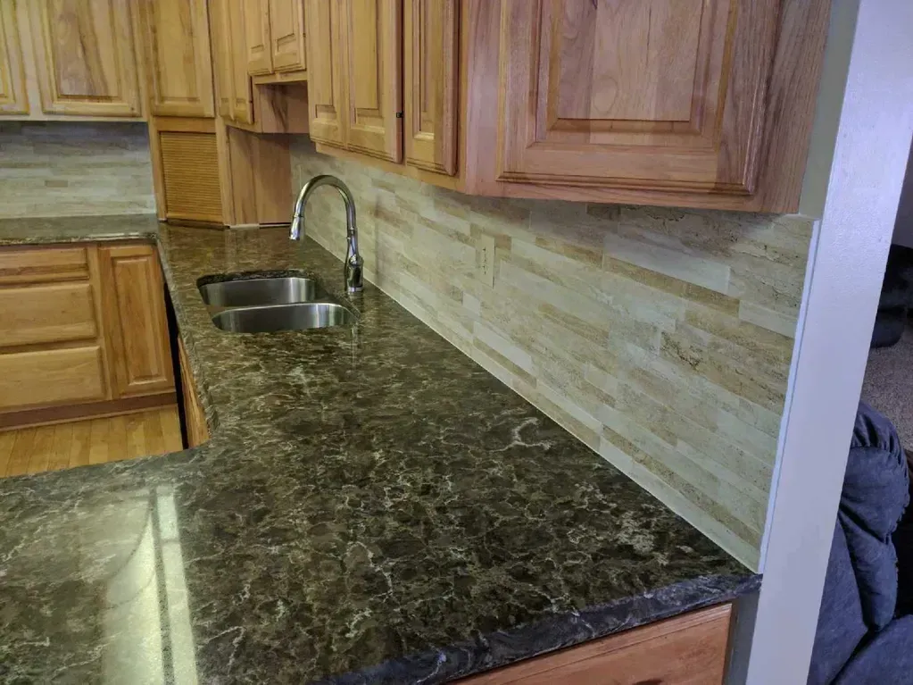 A kitchen view featuring a dark stone countertop, a silver sink faucet, and a textured, light-colored stone backsplash.